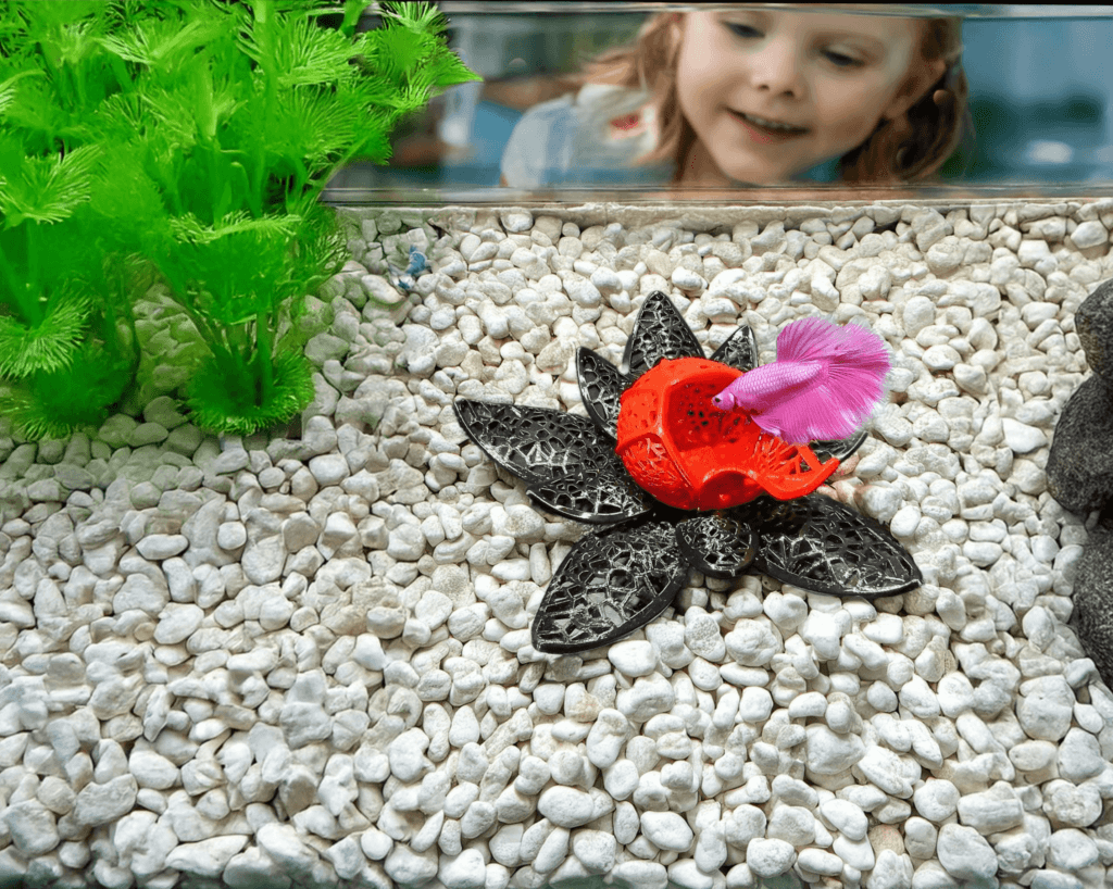 A vibrant, lotus-inspired betta fish cave designed by Aquireef3D, displayed in an aquarium with white gravel and lush greenery, featuring a pink betta exploring the colorful hideaway.