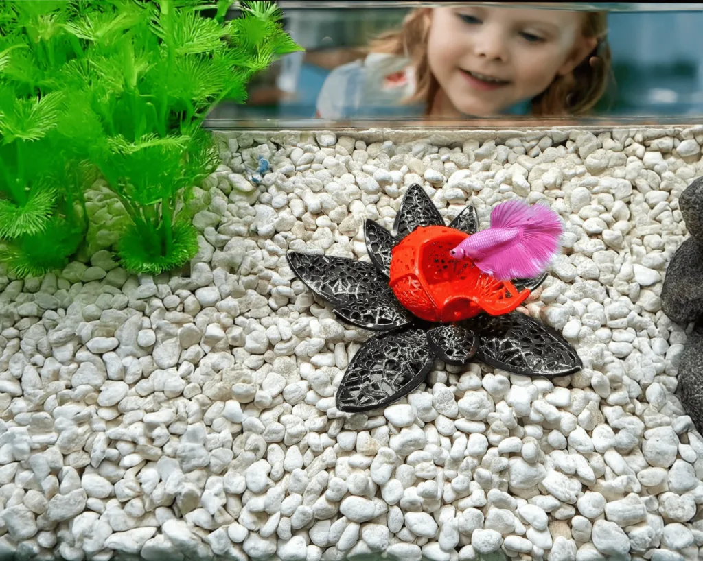 A vibrant, lotus-inspired betta fish cave designed by Aquireef3D, displayed in an aquarium with white gravel and lush greenery, featuring a pink betta exploring the colorful hideaway.